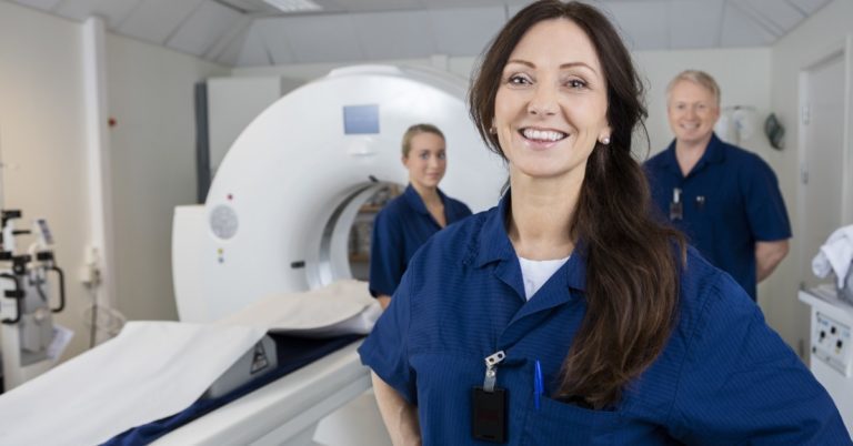 image of a woman with long brown hair in dark blue scrubs standing in front of an MRI machine smiling. there are 2 other radiology technologists in the background smiling