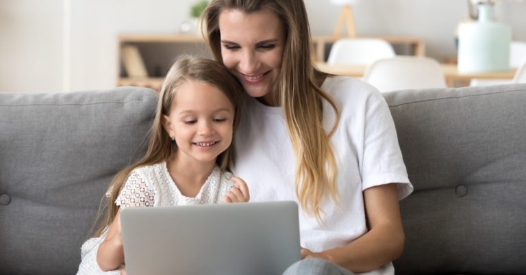 image of a white woman with long hair sitting on a grey couch next to a young girl, both are looking at a laptop in the woman's lap and smiling slightly