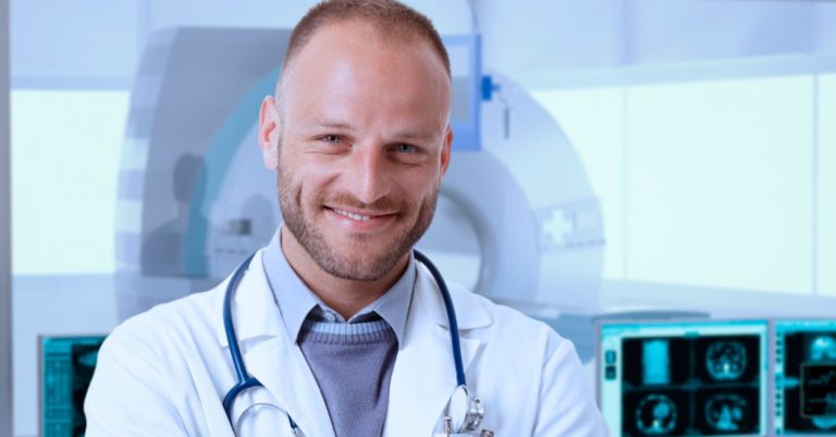 image of a white man in a lab coat and a stethoscope around his neck with arms crossed standing in front of MRI machine smiling at the camera