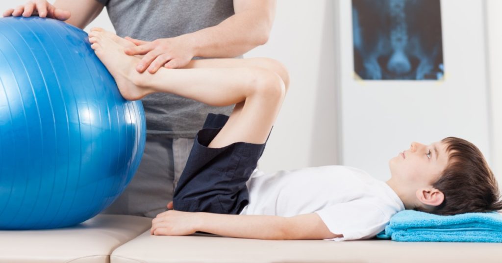 image of school-aged boy laying on exam table with legs bent at 90 degrees and feet up against a sability ball. a man's hands are resting on the boys feet.