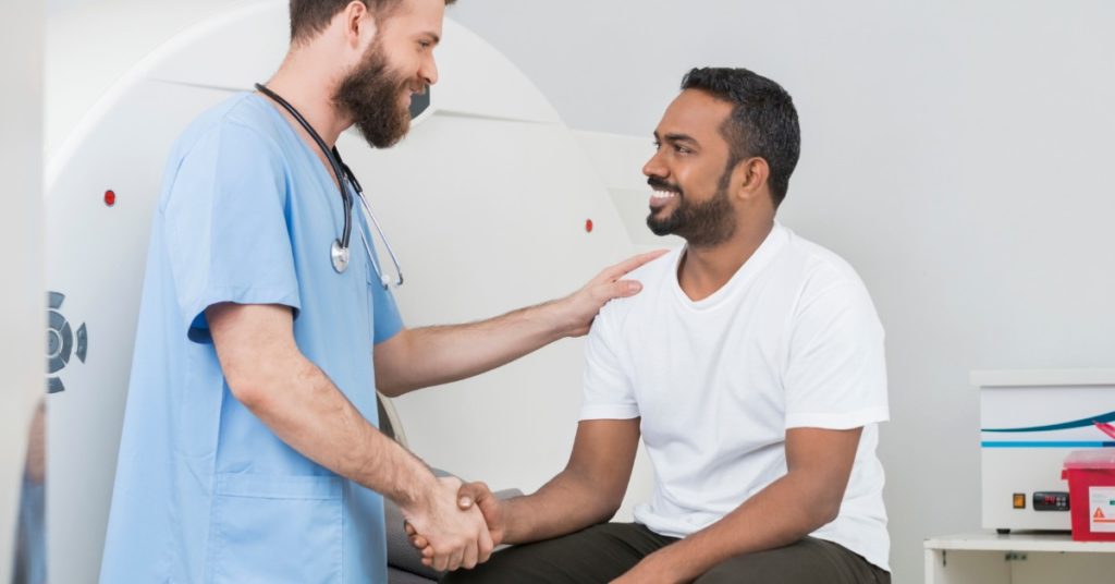 image of a brown man with beard sitting on CT scanner table shaking the hand of a white bearded man in scrubs standing in front of him. both men are smiling