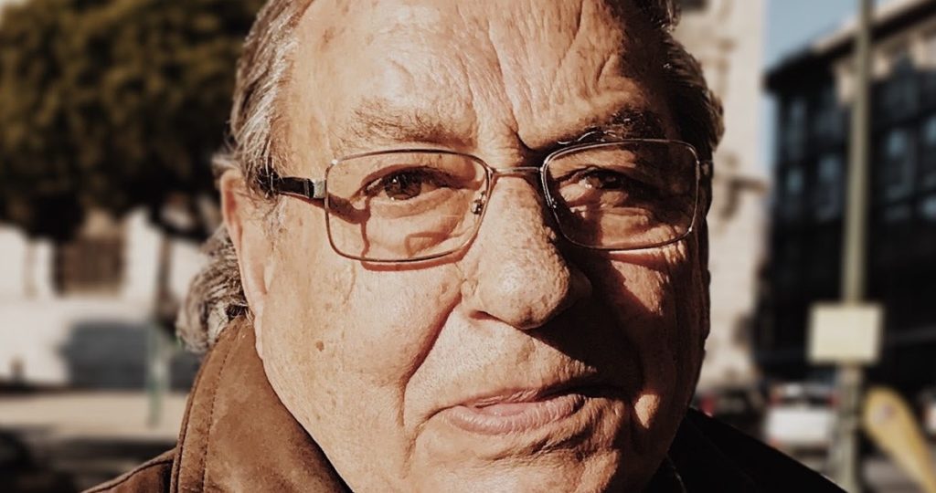 close up image of an older man's face wearing glasses and a jacket, standing outside