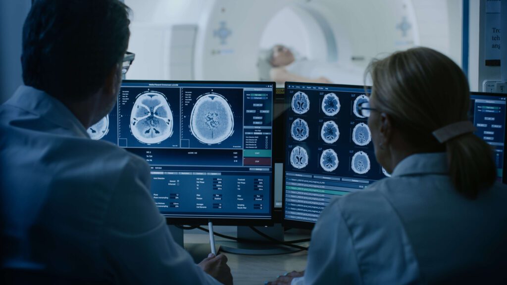 Male and female radiologists reading brain MRI images with a female patient being scanned in a MRI