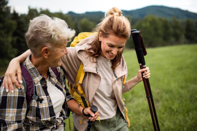 A happy mid adult woman with trekking poles hiking with active senior mother outdoors in nature.