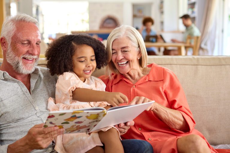 Granddaughter Reading With Grandparents On Sofa At Home With Parents In Background