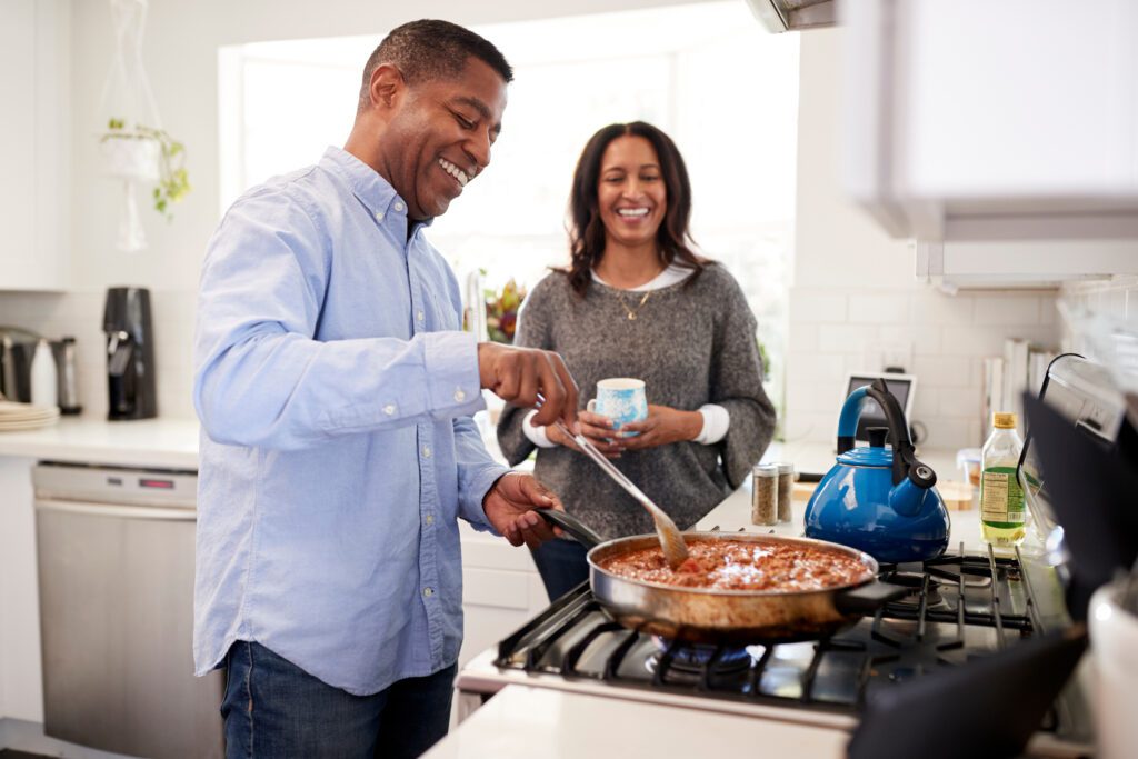 Middle aged man standing in the kitchen cooking with his partner standing beside him, backlit